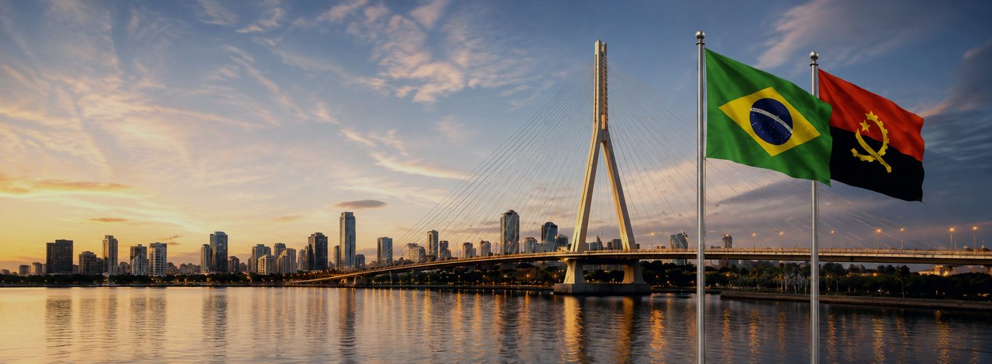 Bandeiras do Brasil e de Angola hasteadas diante de uma ponte estaiada ao entardecer, com a cidade ao fundo.